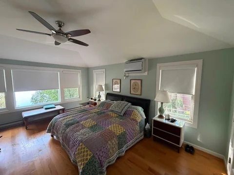 Bright bedroom with green walls, queen bed with colorful patterned bedspread, ceiling fan, hardwood floor, and multiple windows with roller shades
