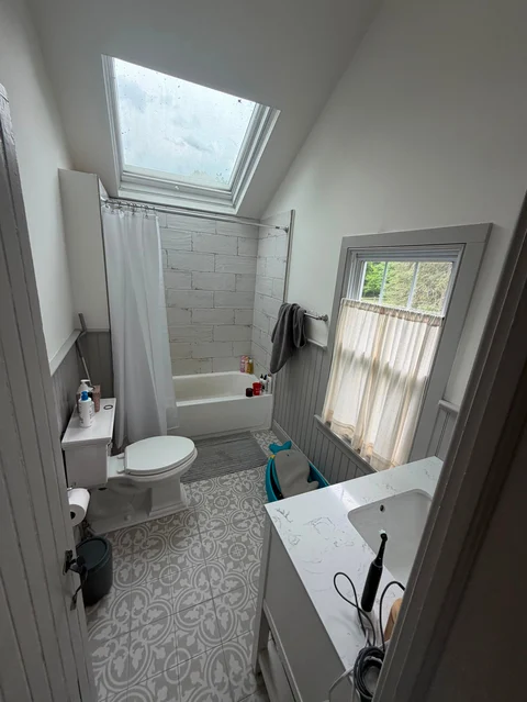 Bright modern bathroom with skylight, white subway tile shower, toilet, vanity sink, patterned floor, and window with natural light