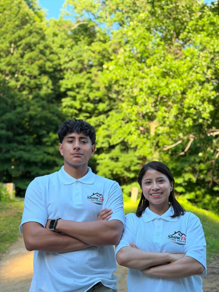 Two young people in matching light blue ShimFit polo shirts standing outdoors with arms crossed, green foliage and clear sky in background